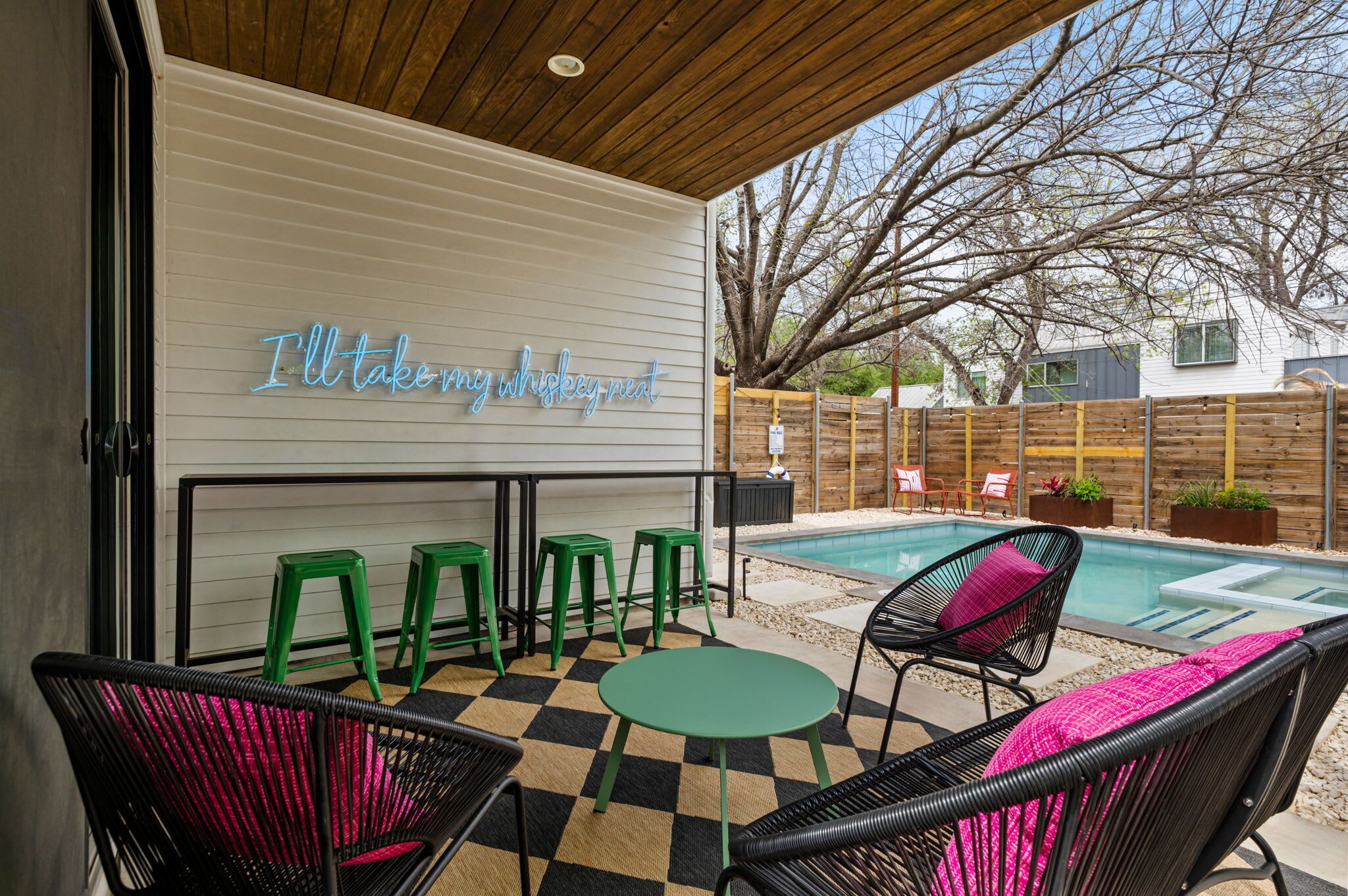 A poolside area with a neon sign, outdoor seating, and a view of a house in the background.