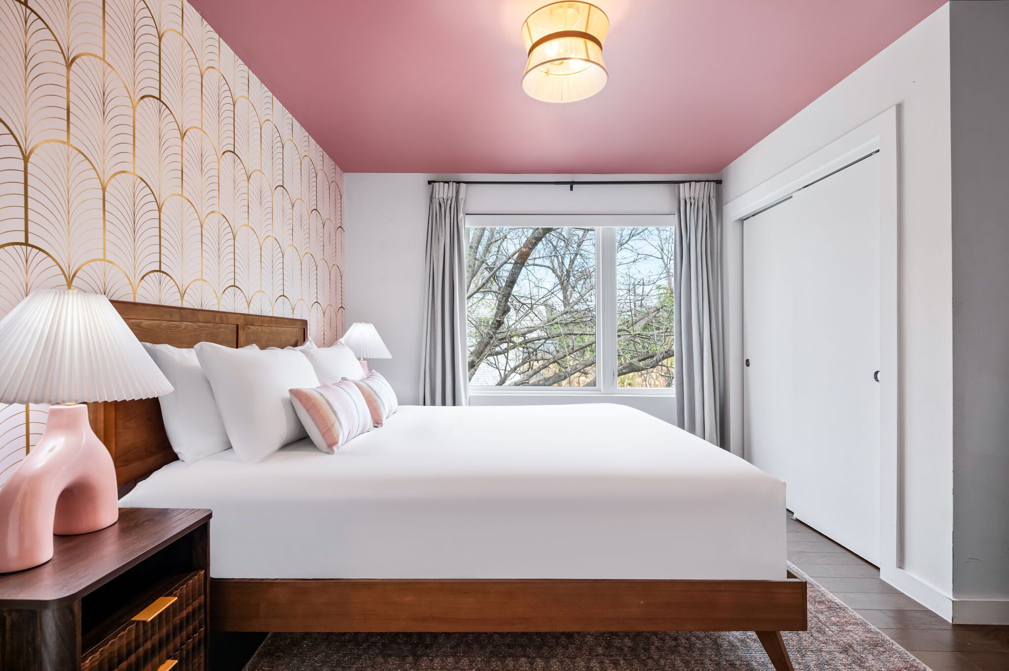 A bedroom with a large bed, a wooden headboard, a nightstand with a lamp, a window with curtains, and a view of trees.
