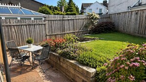Eine Terrasse mit Tisch und Stühlen, ein erhöhter Gartenbeet aus Holz mit Blumen und ein Rasen, der von einem Holzzaun umgeben ist.