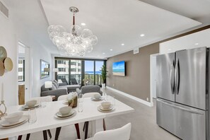 A modern kitchen with a stainless steel refrigerator, a dining area with a white table set for four, and a large window overlooking a balcony with a view of the ocean. - 3BR 2BA Oceanfront Retreat • Pools & Beach Access 706 (Miami Beach)