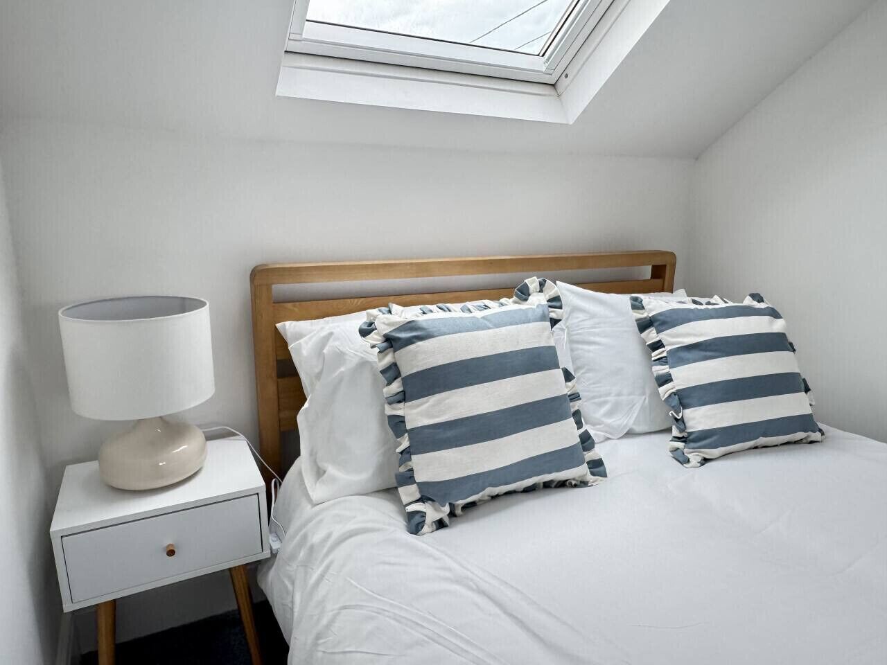 Une chambre avec une lucarne, une tête de lit en bois, une table de chevet avec une lampe et deux coussins à rayures.