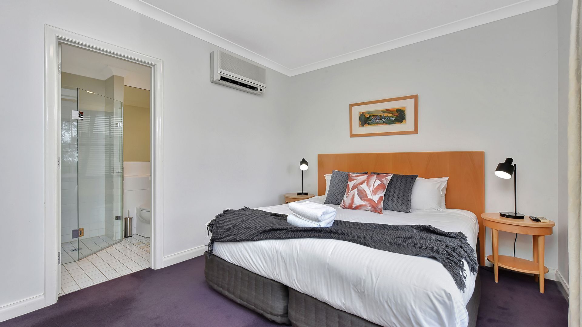 Une chambre d’hôtel comprenant un lit, deux tables de chevet, une commode, un appareil de climatisation, un tableau encadré au mur et une salle de bain avec une cabine de douche en verre.