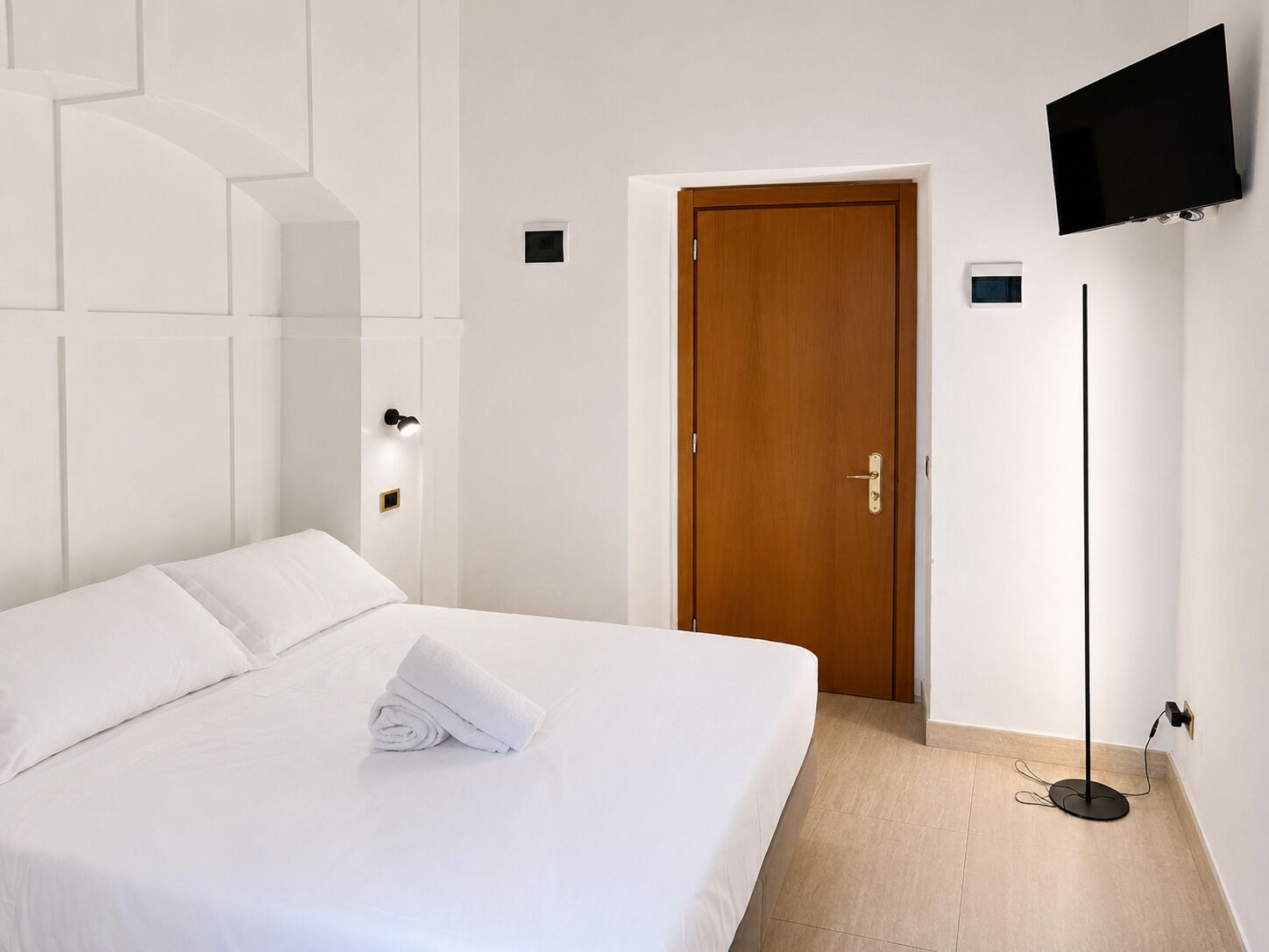 A hotel room with a bed, a wooden door, a flat-screen TV mounted on the wall, and a floor lamp.