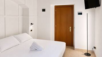 Una camera d'albergo con un letto, una porta in legno, una TV a schermo piatto fissata al muro e una lampada da terra.