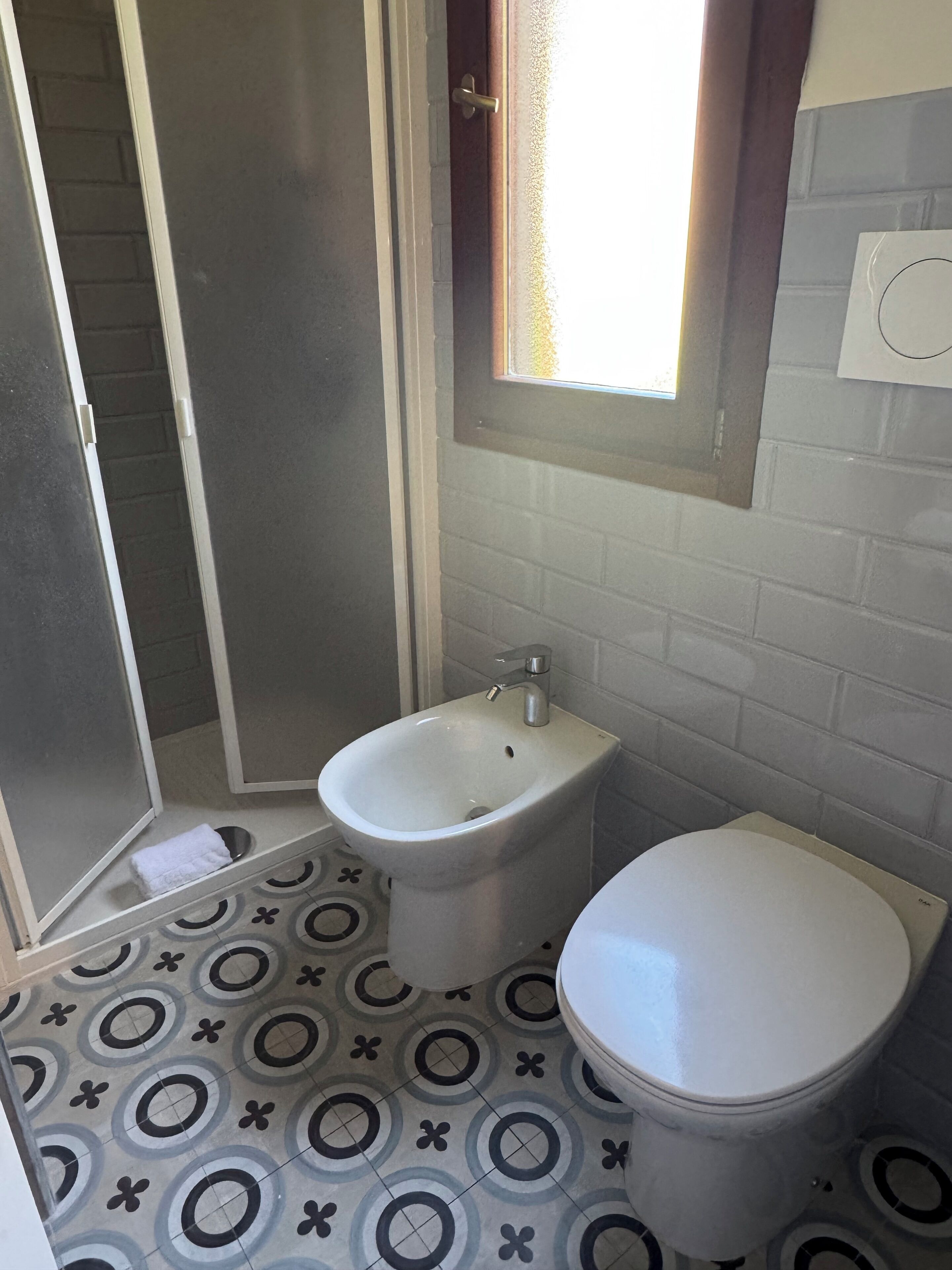 A bathroom with a bidet, toilet, patterned floor, and a window with a view.