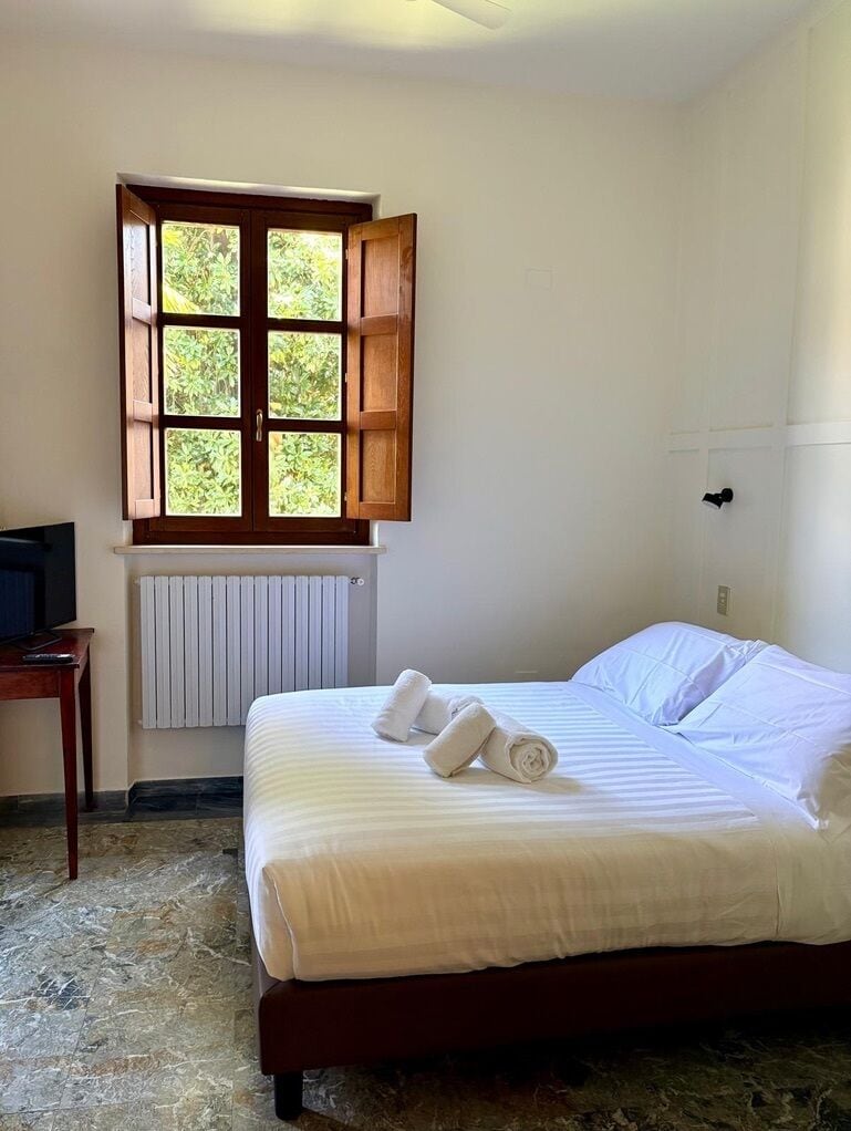 A hotel room with a bed, a window with wooden shutters, a radiator, and a desk with a television.