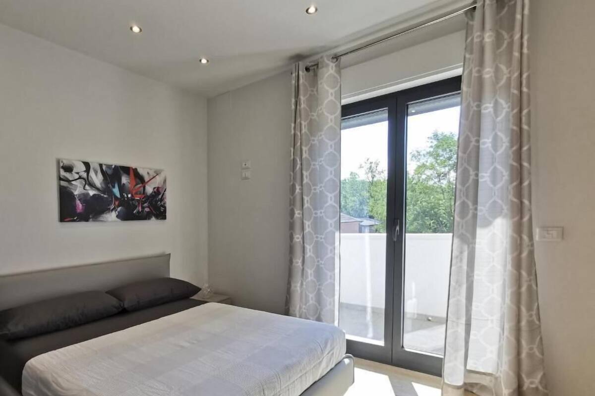 Een slaapkamer met een bed, een schilderij, een raam met gordijnen en uitzicht op groen.