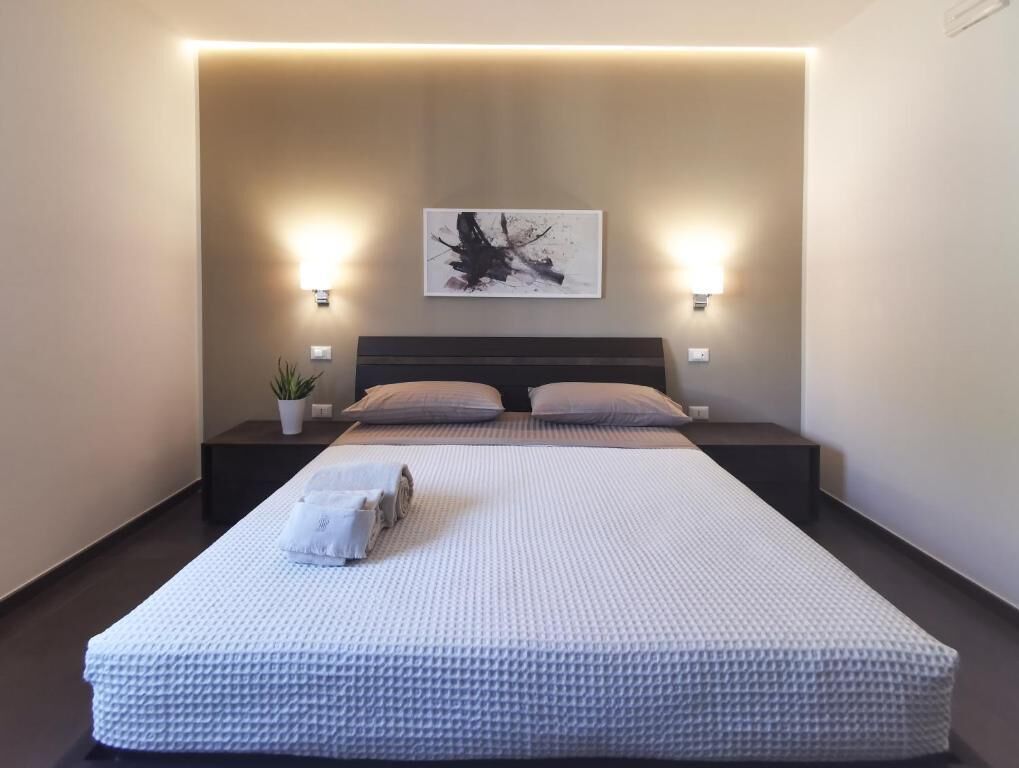 A neatly made bed with a textured white cover, two bedside tables with lamps, a wall-mounted abstract painting, and a potted plant.