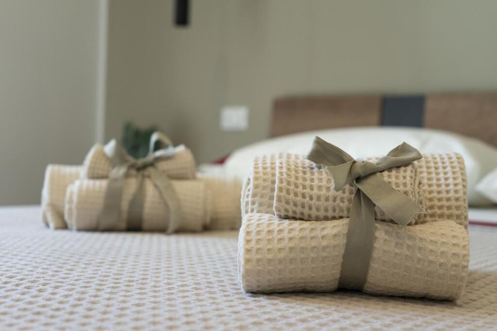 A neatly made bed with a textured white bedspread and two rolled-up towels tied with a beige ribbon.