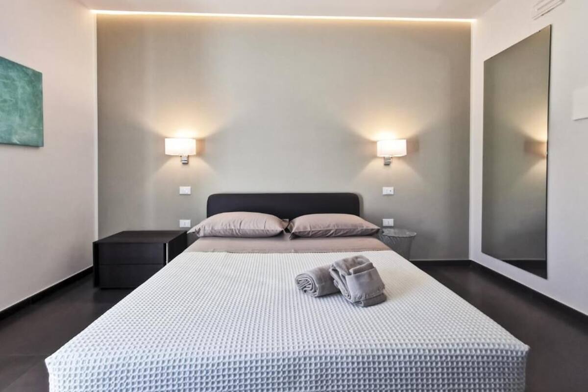Een moderne hotelkamer met een groot bed, twee bedlampen, een klein nachtkastje en een grote spiegel aan de muur.