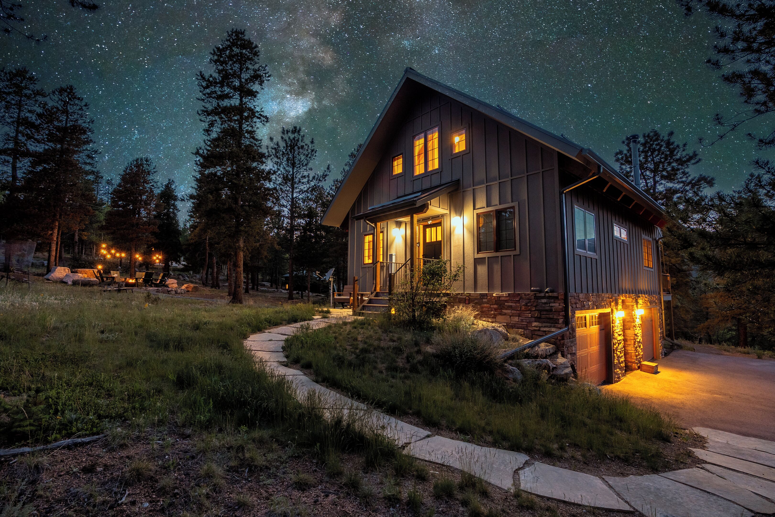 A two-story cabin with a stone foundation, a porch with outdoor seating, and a clear view of the night sky.