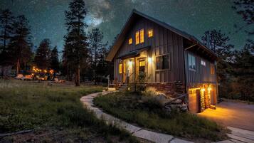 A two-story cabin with a stone foundation, a porch with outdoor seating, and a clear view of the night sky.