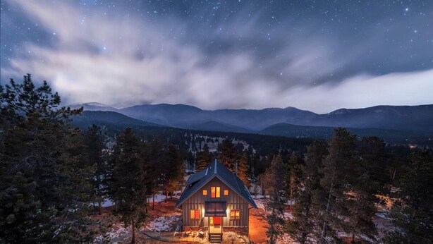 A cabin with a stone exterior, surrounded by snow-covered ground and trees, under a starry night sky.