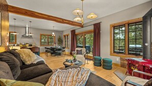 A cozy living room with a fireplace, wooden beams, large windows, and a dining area.