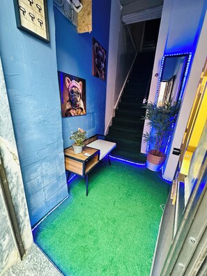 Ein schmaler Flur mit künstlichem Grünteppich, einer kleinen Holzbank mit Topfpflanze und einer Treppe mit blauer LED-Beleuchtung.