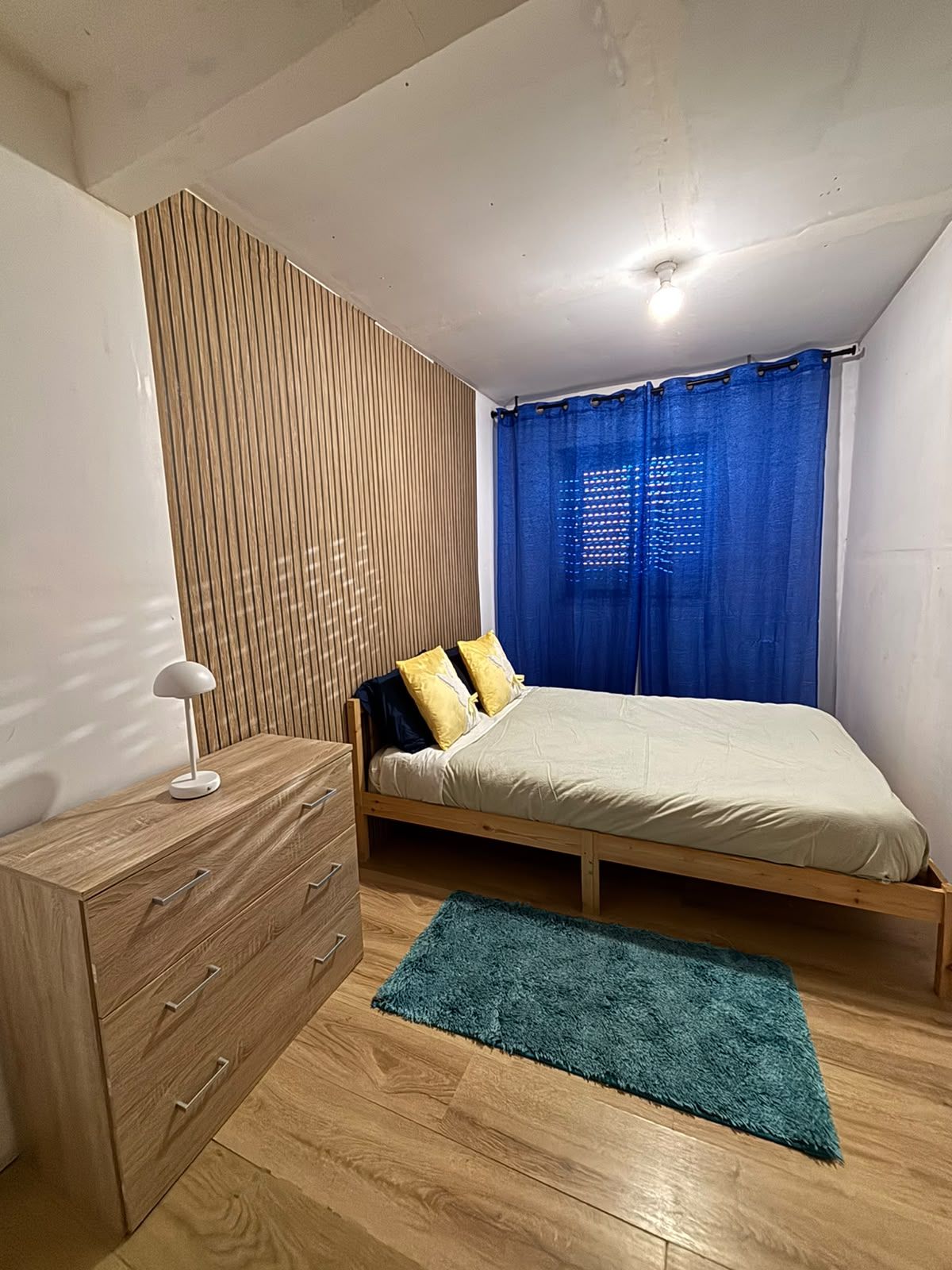 Une chambre à coucher avec un lit en bois, une table de chevet, une commode, un rideau bleu et un luminaire au plafond.
