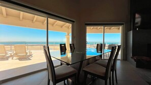 A room with a glass table and chairs, a TV, and a view of the beach. - The Beach House | 3BR Beachfront Home | Sleeps 10 (Puerto Peñasco)
