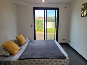 Un dormitorio con cama, dos almohadas, una ventana con vista a follaje y una lámpara de pared.