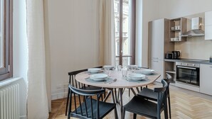 Uma sala de jantar com mesa redonda posta para quatro pessoas, cozinha com eletrodomésticos modernos e janela com vista para prédios.