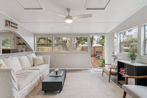 A spacious living area with a white sofa, a coffee table, and a TV stand. There is a ceiling fan and large windows providing natural light. - Game Room | Hot Tub | Near Beach & Pier | Fire Pit | Bunk Beds | Pet Friendly (St. Simons Island)