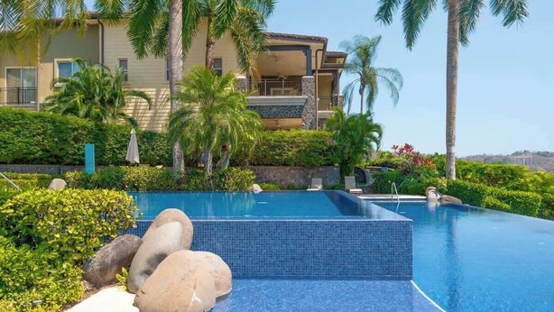 A pool with clear blue water, surrounded by rocks and palm trees, with a building in the background. - Oceanview Condo with Private Pool in Los Sueños (Herradura)