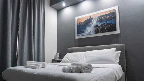 A neatly made bed with rolled towels, a gray headboard, a framed coastal photograph on the wall, and a nightstand with a lamp. - Deluxe Double Room 101 (Catania)