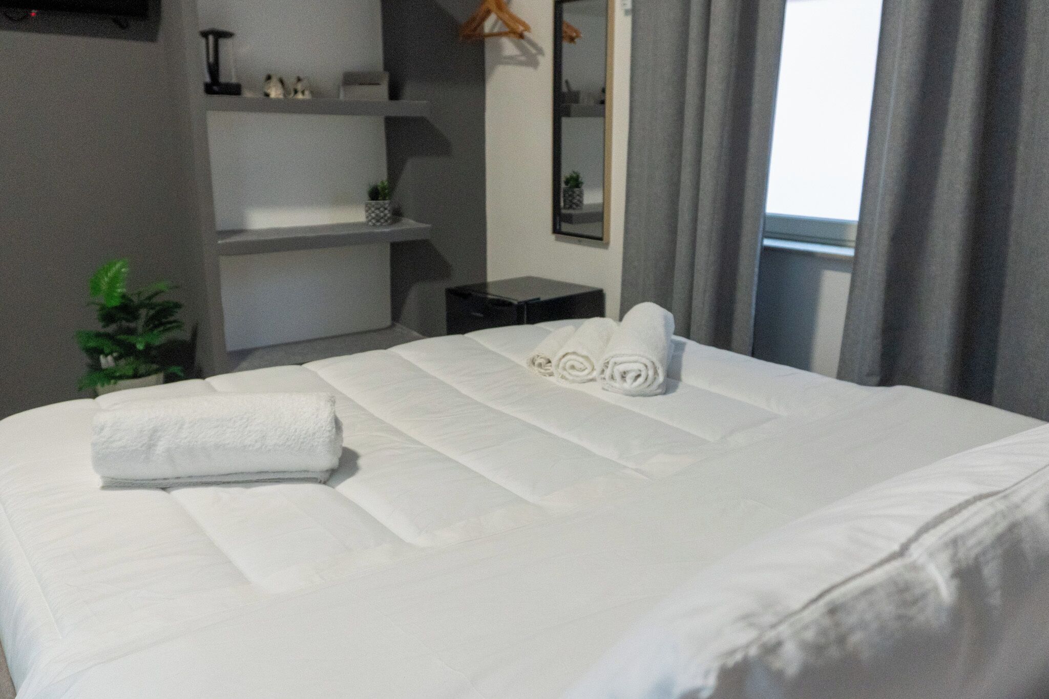 A neatly made bed with white linens and rolled towels, a small bedside table, a mirror, and a potted plant.