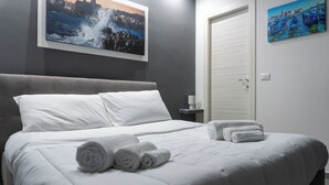 A neatly made bed with white linens and rolled towels, a painting of a coastal scene, and a closed door. - Deluxe Double Room 101 (Catania)