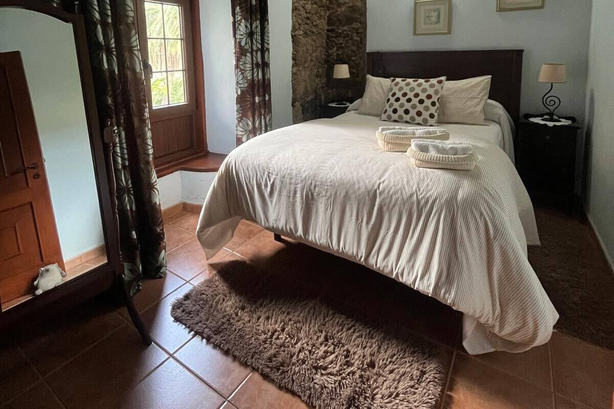 A bedroom with a bed, a nightstand, a lamp, a window with curtains, and a stone wall.