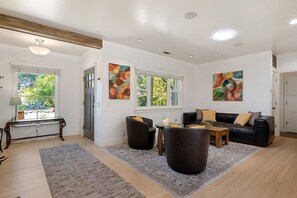 A spacious living room with a sofa, armchairs, a coffee table, and a rug. There are two windows with curtains, a ceiling light, and abstract paintings on the walls.