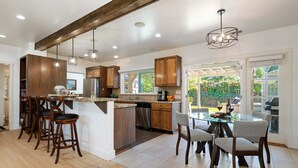 A modern kitchen with wooden cabinets, a central island with bar stools, and a dining area with a glass table and chairs. - NEW Steps to the Sand on Mesa Lane 3 BD 2BA with Jacuzzi! (Santa Barbara)