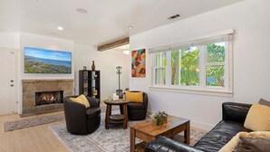 A living room with a fireplace, a large window, and a view of greenery outside. - NEW Steps to the Sand on Mesa Lane 3 BD 2BA with Jacuzzi! (Santa Barbara)