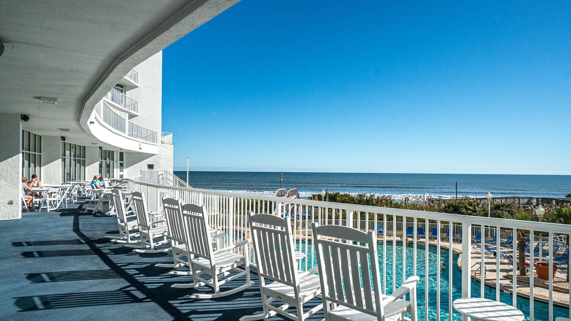 A balcony with white rocking chairs and a pool, overlooking the ocean.