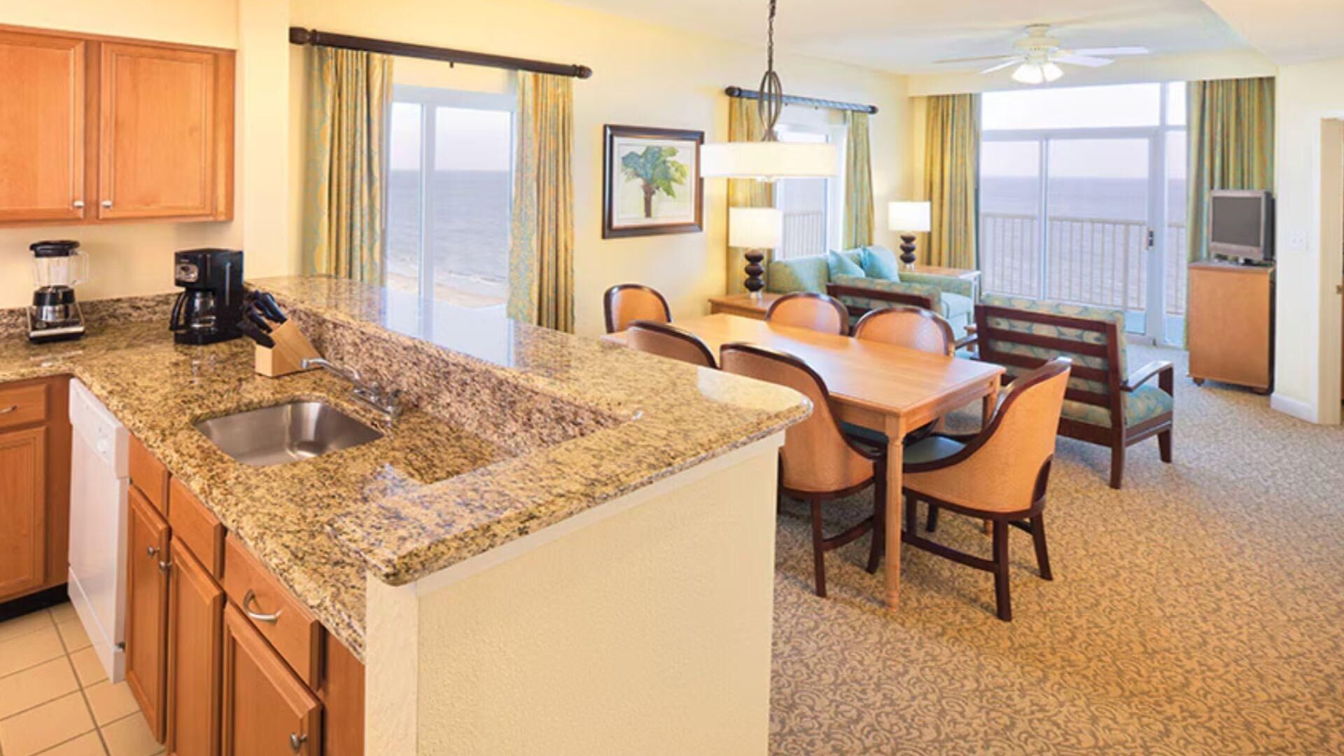 A hotel room with a kitchen area, dining table, chairs, and a balcony with a view.