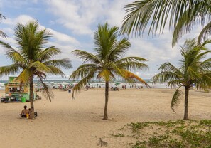 A beach with palm trees, a food stall, and a sandy shore. - Apartamento Beira Mar com Varanda e Churrasqueira (Praia Grande)