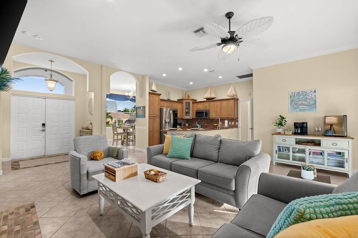 A spacious living area with a grey sofa, a white coffee table, and a grey armchair. There is a kitchen in the background with wooden cabinets and a ceiling fan.