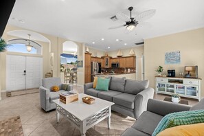 A spacious living area with a grey sofa, a white coffee table, and a grey armchair. There is a kitchen in the background with wooden cabinets and a ceiling fan.