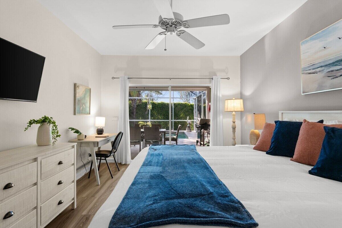 Et soveværelse med seng, skrivebord, stol, ventilator i loftet og udsigt gennem en glasdør.