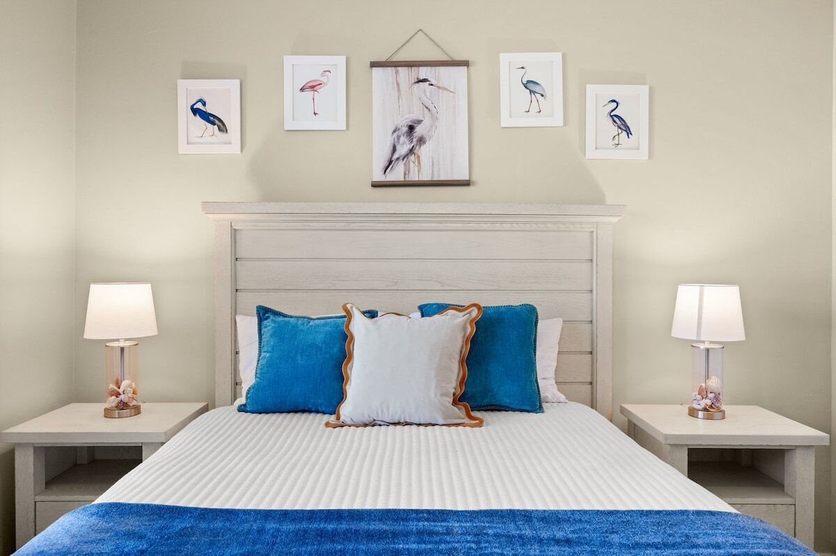 Une chambre d’hôtel comprenant un lit, deux tables de chevet, une petite table, une tête de lit et quatre œuvres d’art encadrées accrochées au mur.