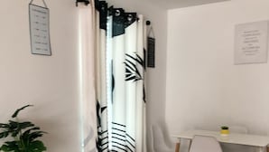 Una habitación con una mesa de comedor, sillas, una planta en maceta, una cortina con estampado de hojas y un letrero en la pared.