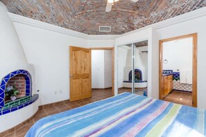 Ein Schlafzimmer mit Ziegeldecke, Kamin, einem Bett mit gestreifter Bettdecke und einem Badezimmer mit gefliestem Duschbereich.
