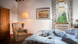 A bedroom with a bed, a chair, a table with a tray, a lamp, a painting, and a window with curtains. - Signorelli (Umbria)
