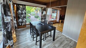 A dining area with a wooden table and chairs, a view of a garden through large windows, and a ceiling with wooden beams. - Sandmo Holiday Home 11 (Ålands landsbygd)