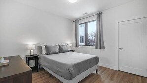 Um quarto com cama, mesas de cabeceira, janela com cortinas, porta e luminária no teto.