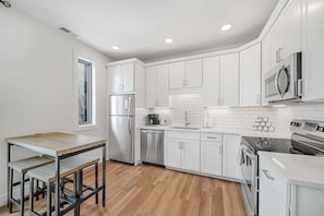 A modern kitchen with white cabinets, stainless steel appliances, and a wooden dining table. - Downtown Condo | Walk to Bars, Beach + Food (Traverse City)