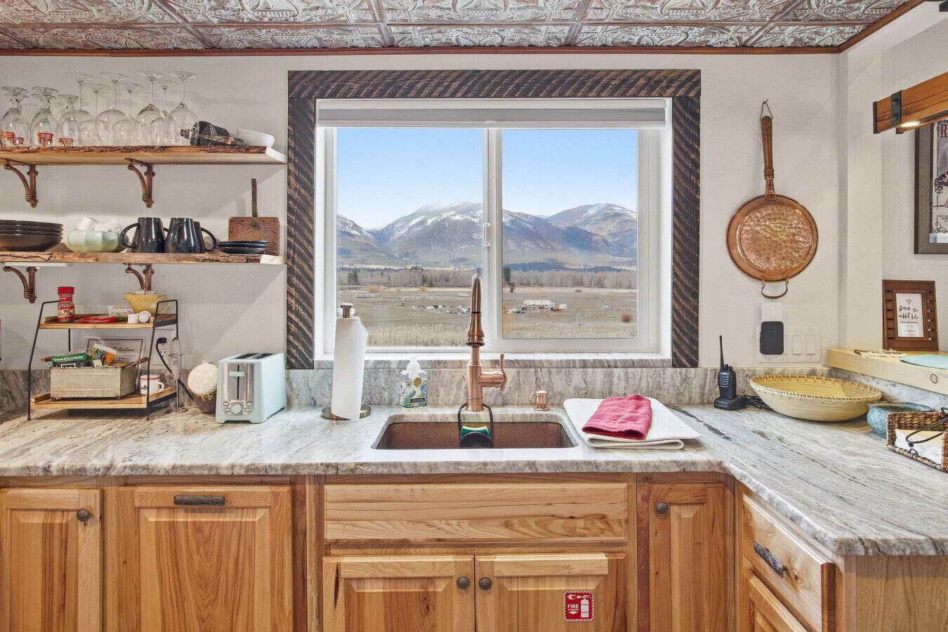 Cucina con mobili in legno, piano di lavoro in marmo, una finestra vista montagna e una pentola di rame appesa.