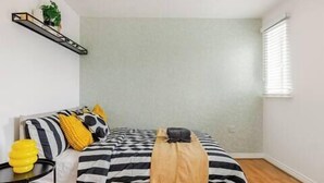 Un dormitorio con cama, mesita de noche, lámpara, planta y ventana.
