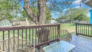 A wooden deck with wicker furniture and a glass table, surrounded by a large tree and a grassy yard. - Awake to Adventure, then Recharge and Relax in Historic Georgetown, Texas! (Georgetown)