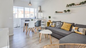 Un salon moderne avec un canapé gris, une table basse blanche et des chaises noires. On y trouve également un coin cuisine avec des meubles blancs et une table à manger. La pièce est dotée d’une grande fenêtre qui laisse entrer la lumière naturelle.