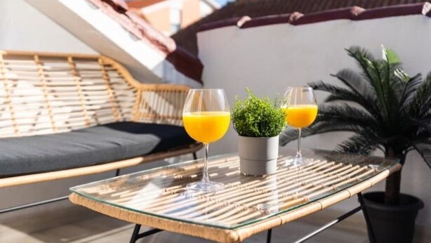 Une table en verre avec deux verres de jus d’orange, une petite plante en pot et une chaise en osier avec un coussin.